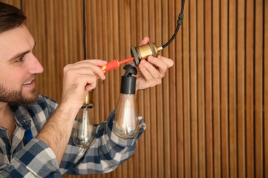 Electrician with screwdriver repairing ceiling lamp against wooden background. Space for text