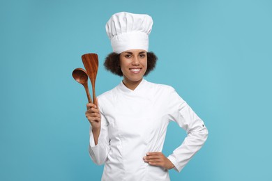 Happy female chef in uniform holding spoon and spatula on light blue background