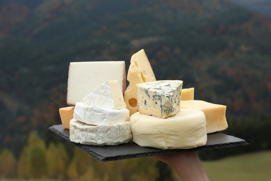 Photo of Woman holding slate plate with different types of delicious cheeses against mountain landscape, closeup