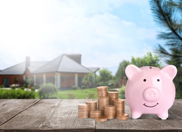 Piggy bank and stacked coins on wooden surface and blurred view of beautiful house, space for text. Mortgage concept