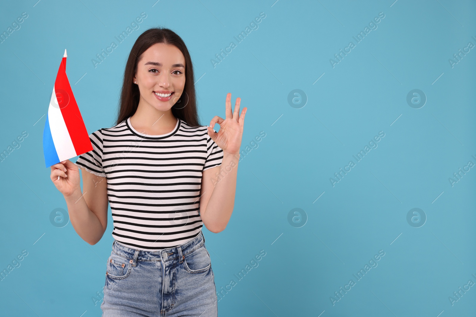 Photo of Young woman with flag of Netherlands showing ok gesture on light blue background, space for text