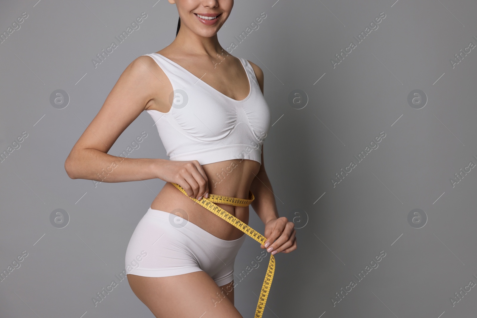 Photo of Young woman measuring waist with tape on grey background, closeup. Space for text