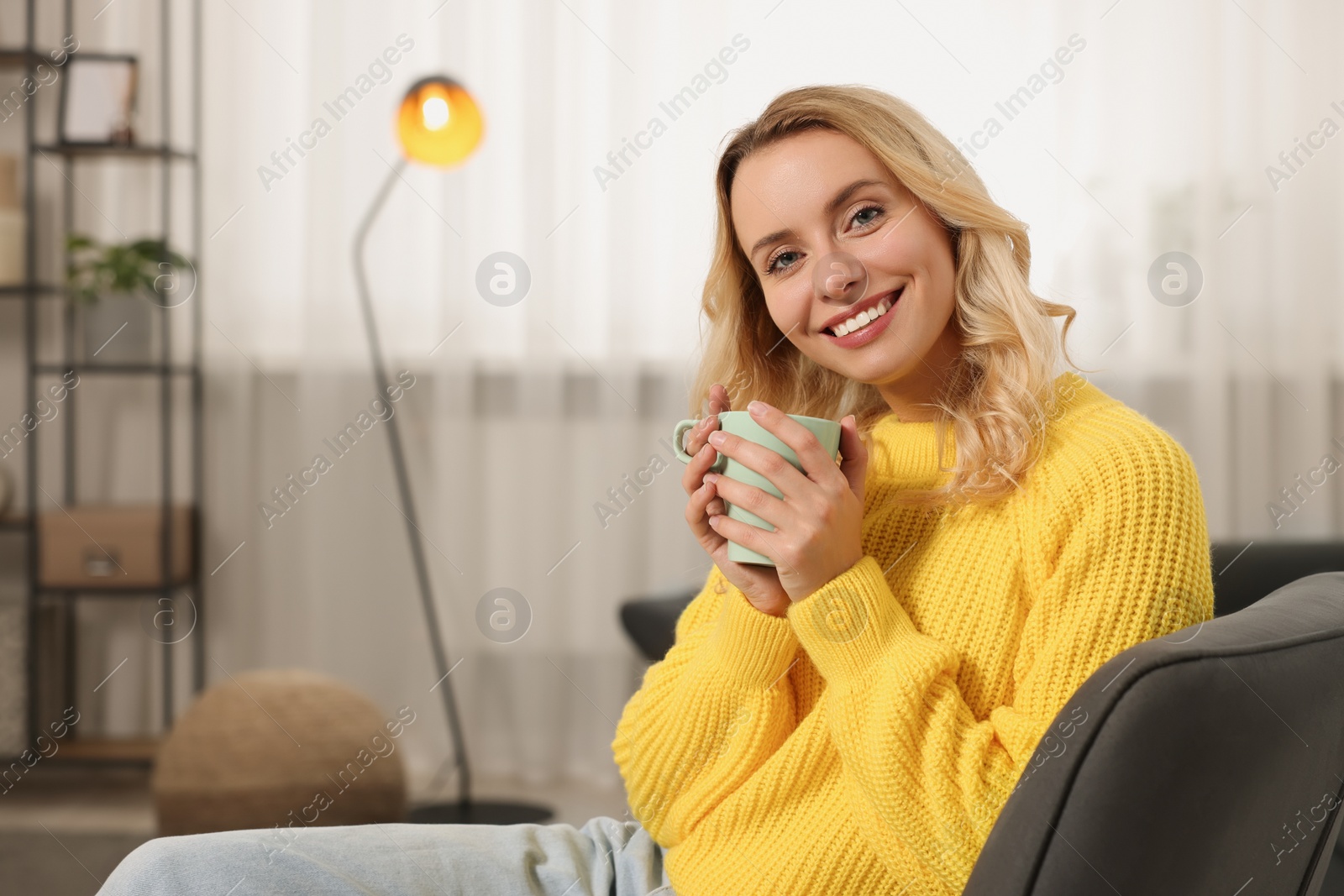 Photo of Happy woman in stylish warm sweater holding cup of drink at home. Space for text