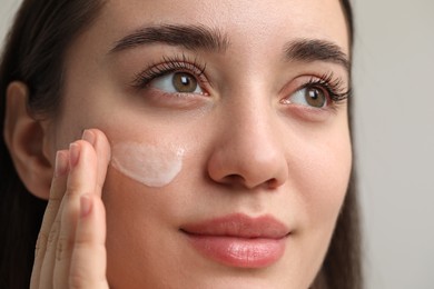 Young woman with dry skin applying cream onto her face on blurred background, closeup