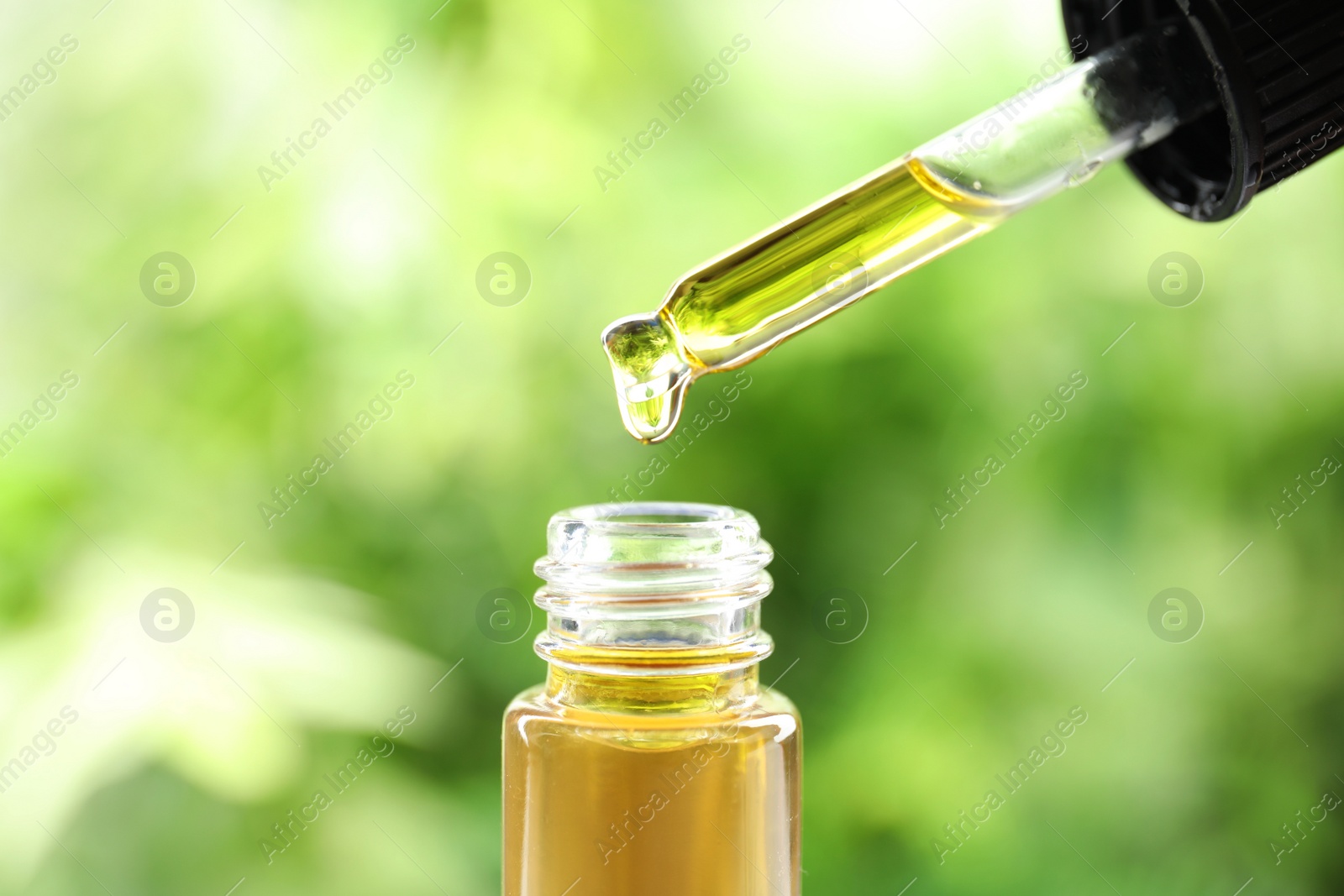 Photo of Dripping essential oil from pipette into glass bottle against blurred green background, closeup