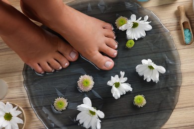 Photo of Woman soaking her feet in plate with water and flowers on wooden floor, top view. Pedicure procedure