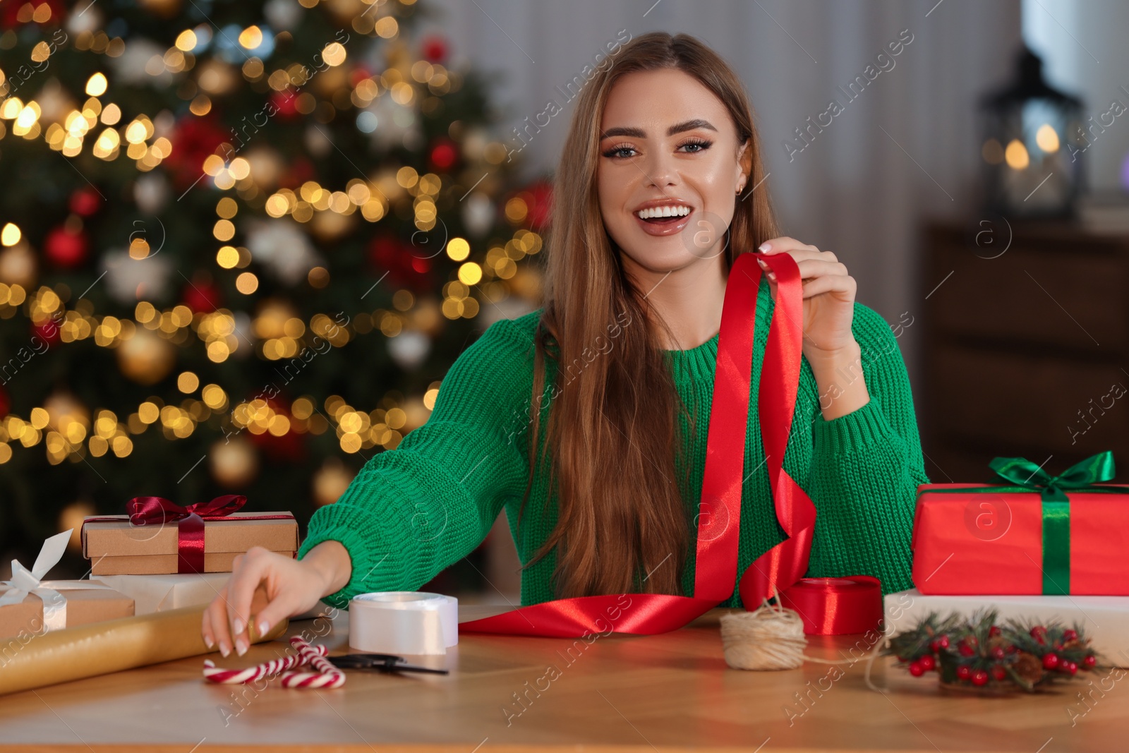 Photo of Beautiful young woman with red ribbon at table indoors. Decorating Christmas gift