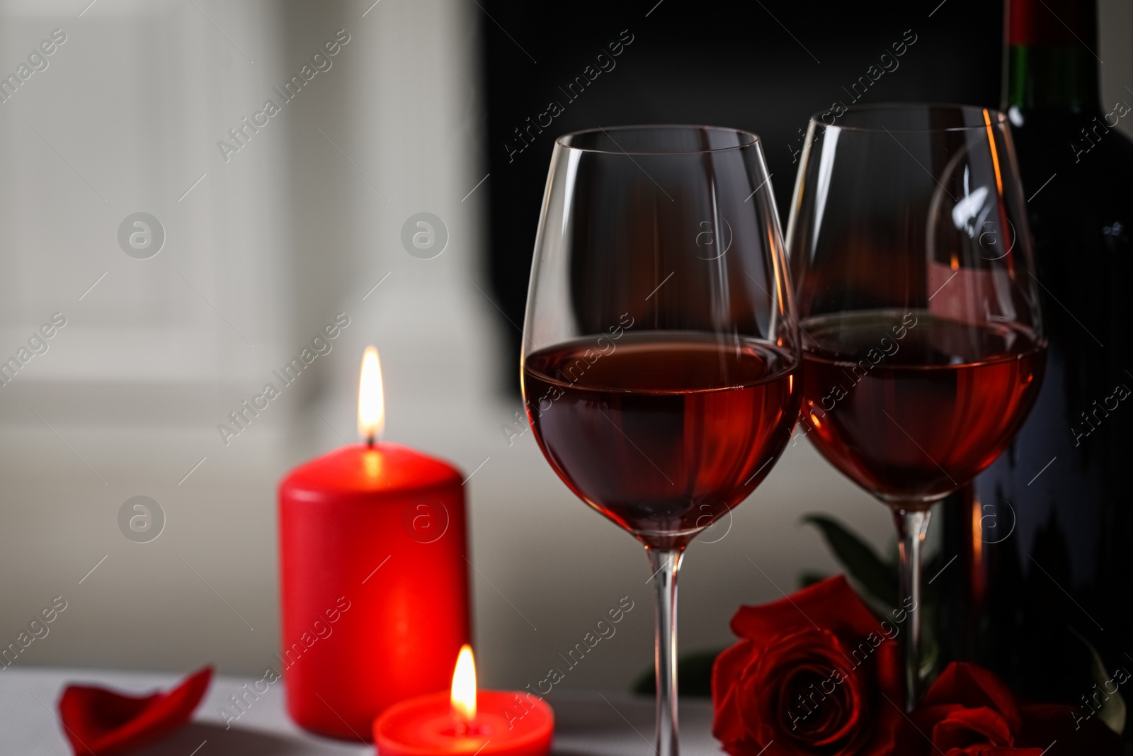 Photo of Glasses of red wine, burning candles and rose flowers against blurred background, space for text. Romantic atmosphere