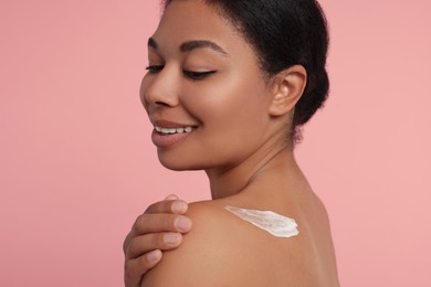 Young woman applying body cream onto back on pink background