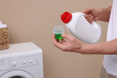 Photo of Man pouring fabric softener from bottle into cap near washing machine indoors, closeup