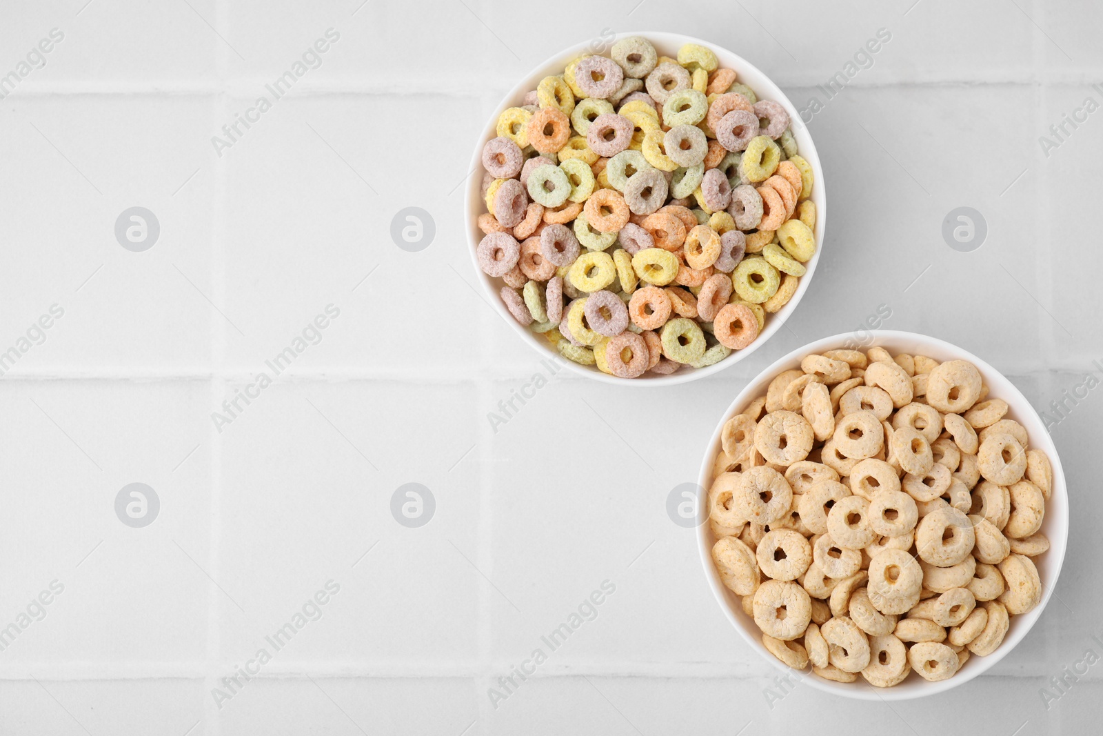 Photo of Tasty cereal rings in bowls on white tiled table, flat lay. Space for text