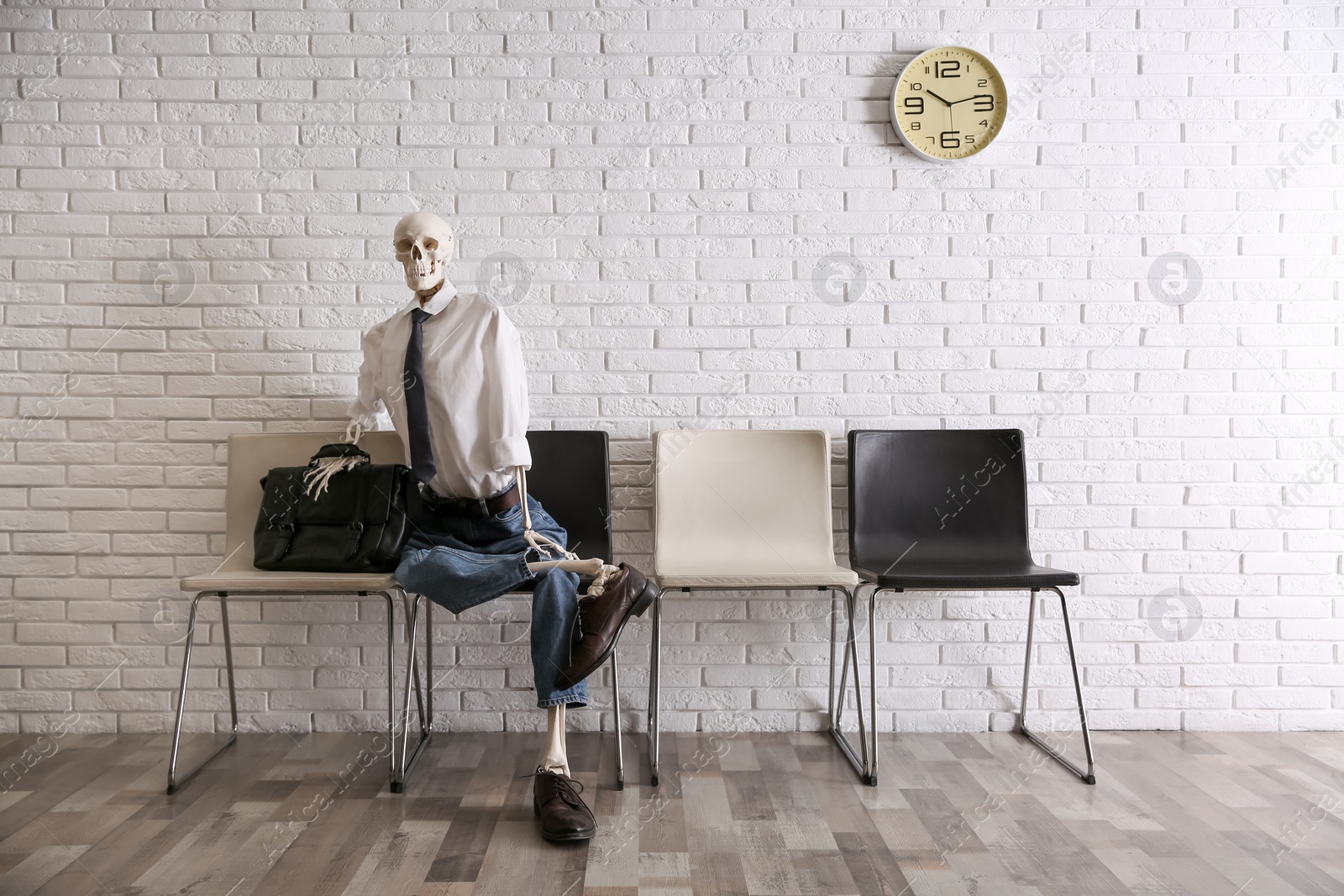 Photo of Human skeleton in office wear sitting on chair near brick wall indoors