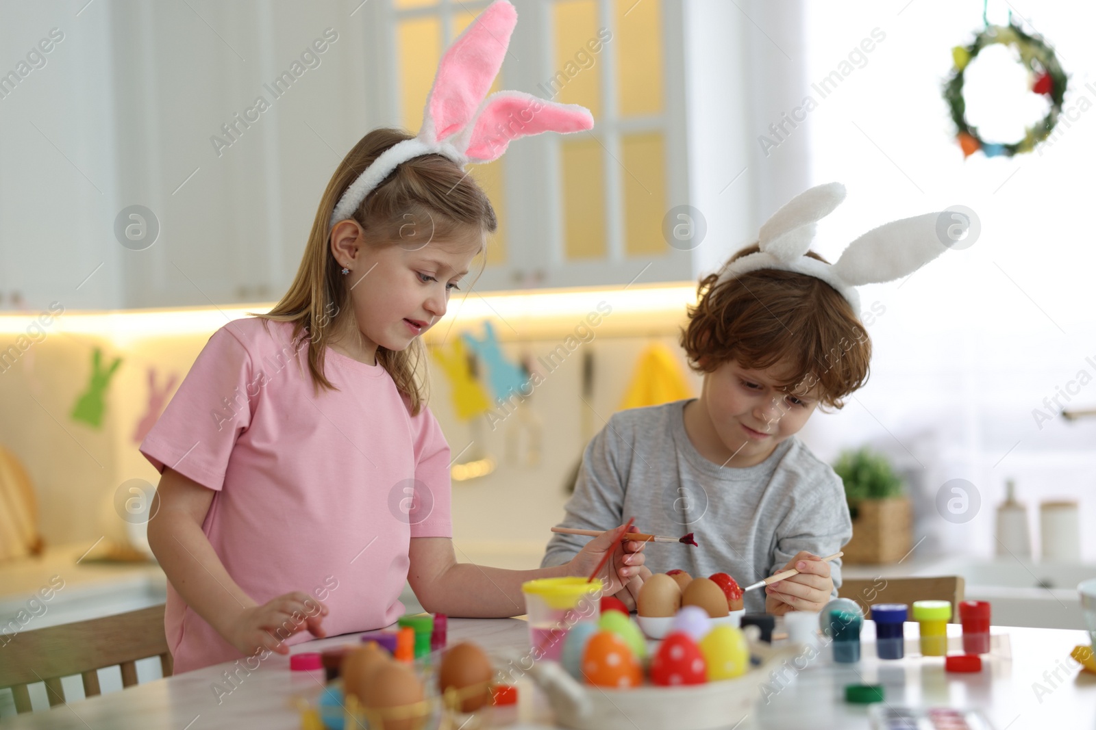 Photo of Easter celebration. Cute children with bunny ears painting eggs at table in kitchen