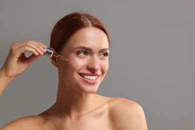 Photo of Beautiful young woman applying cosmetic serum onto her face on grey background, space for text