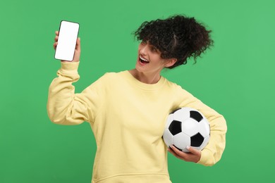 Photo of Happy fan holding soccer ball and showing smartphone on green background