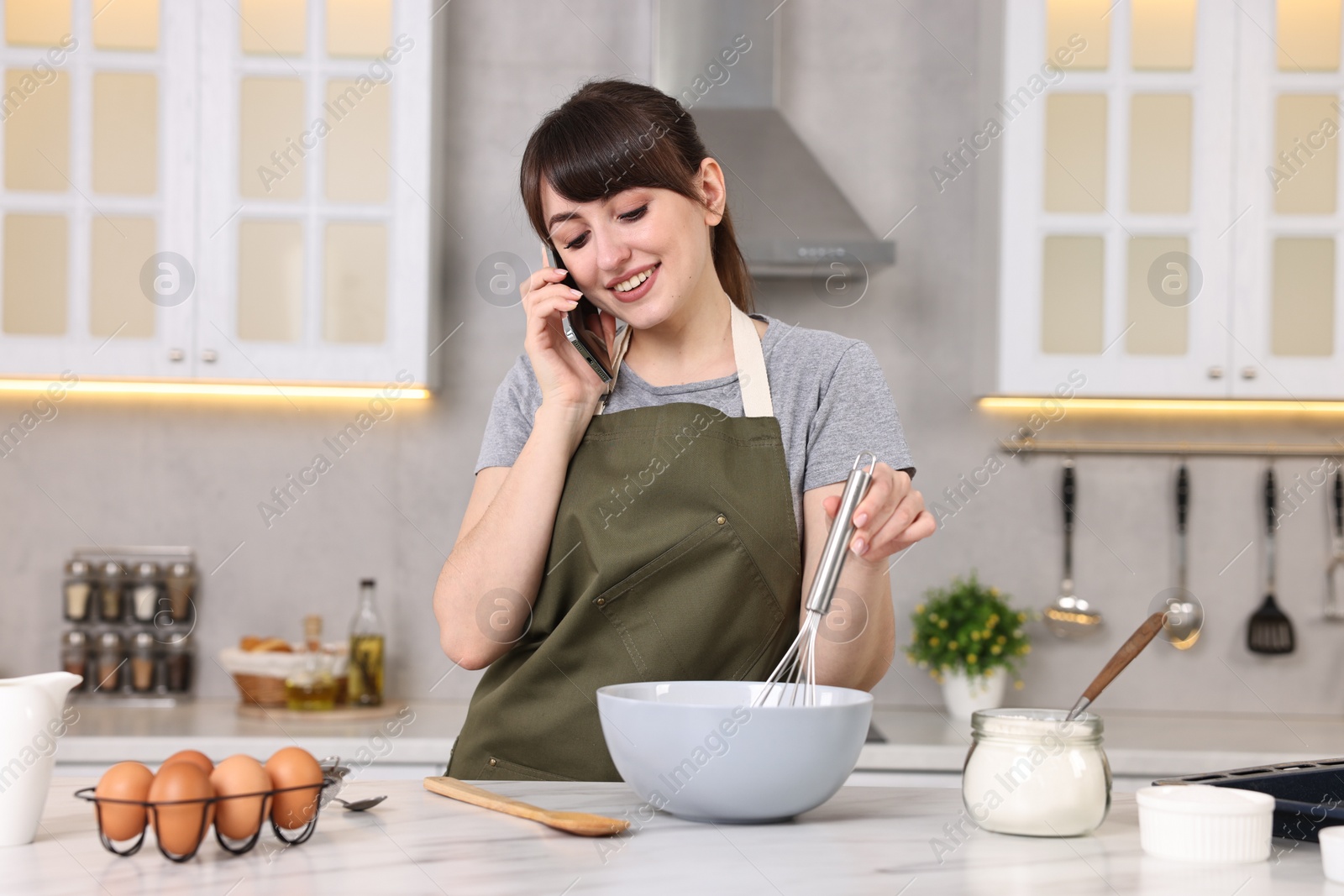 Photo of Happy young housewife talking on smartphone while cooking at white marble table in kitchen