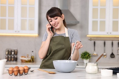Photo of Happy young housewife talking on smartphone while cooking at white marble table in kitchen