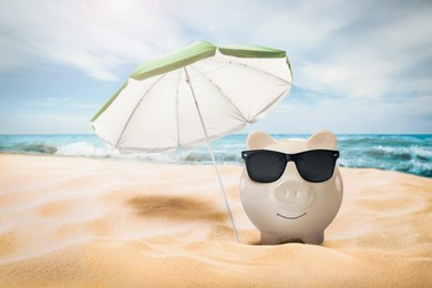 Image of Vacation savings. Piggy bank with sunglasses and umbrella on sandy beach near sea