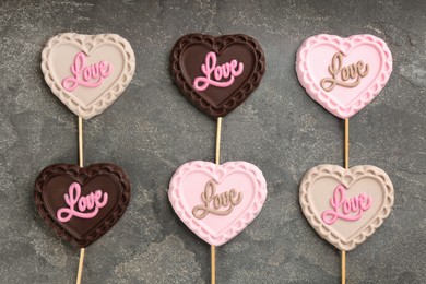 Photo of Different chocolate heart shaped lollipops with word Love on grey table, flat lay