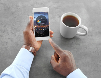 Image of African-American man using weather forecast app on smartphone at table, closeup