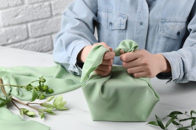 Photo of Furoshiki technique. Woman wrapping gift in green fabric at white table, closeup