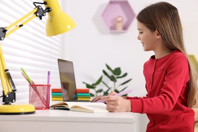 Photo of E-learning. Cute girl taking notes during online lesson at table indoors