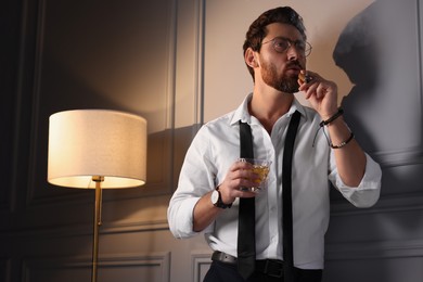 Photo of Handsome man with glass of whiskey smoking cigar at home, low angle view