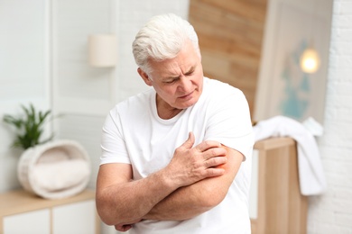 Senior man scratching arm indoors. Allergy symptom