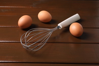Photo of Metal whisk and raw eggs on wooden table, closeup