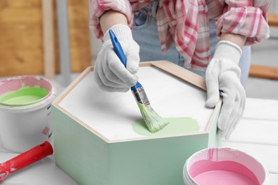 Photo of Woman painting honeycomb shaped shelf with brush at white table indoors, closeup