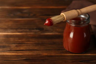 Photo of Tasty barbecue sauce in glass jar and brush on wooden table, closeup. Space for text