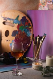 Photo of Glass of tasty wine, brushes with colorful paints and burning candle on light gray table