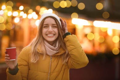 Photo of Happy young woman with cup of drink at winter fair in evening. Christmas celebration