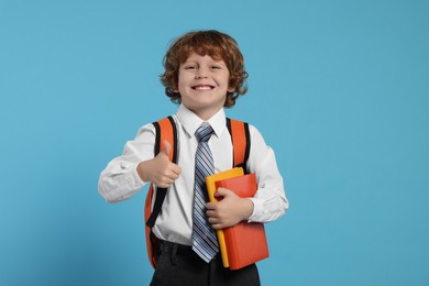 Photo of Happy schoolboy with backpack and books showing thumb up gesture on light blue background