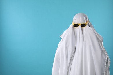 Stylish ghost. Person covered with white sheet in sunglasses on light blue background, space for text