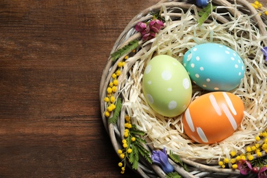 Photo of Wicker nest with painted Easter eggs and flowers on wooden table, top view. Space for text