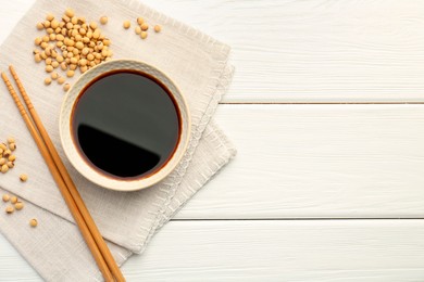 Photo of Tasty soy sauce in bowl, chopsticks and soybeans on white wooden table, top view. Space for text