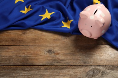 Photo of Pink piggy bank and flag of European Union on wooden table, above view. Space for text