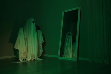 Photo of Creepy ghost. Woman covered with sheet looking at mirror in green light