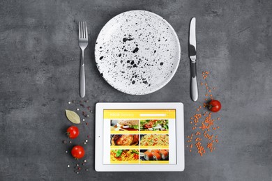 Photo of Modern tablet with open page for online food ordering, scattered tomatoes, spices, plate and cutlery on black table, flat lay. Concept of delivery service