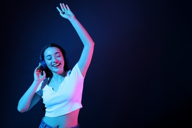 Happy woman in headphones listening music and dancing in neon lights against dark blue background. Space for text