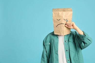 Photo of Woman wearing paper bag with drawn sad face on light blue background. Space for text