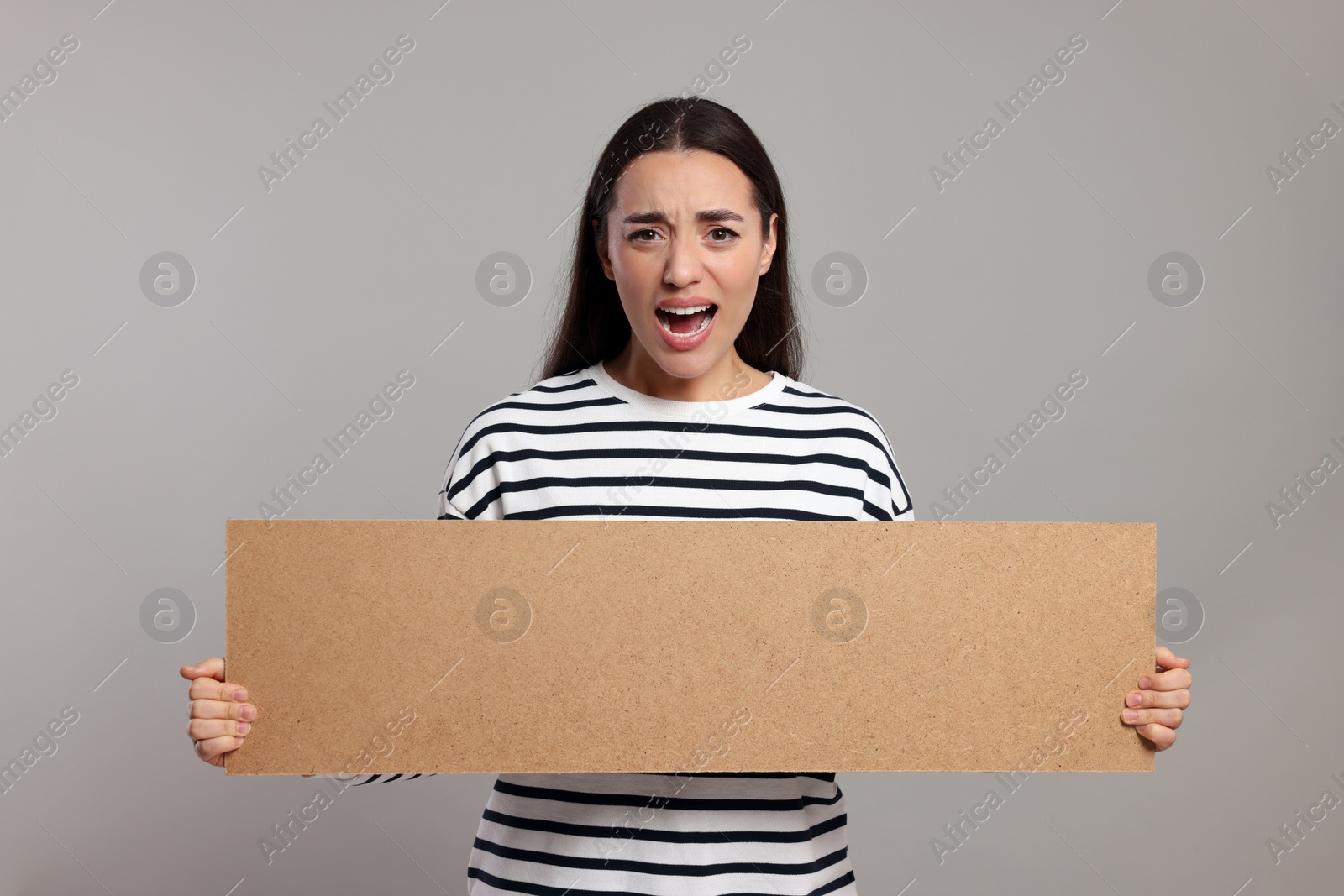 Photo of Angry young woman holding blank cardboard banner on light grey background, space for text
