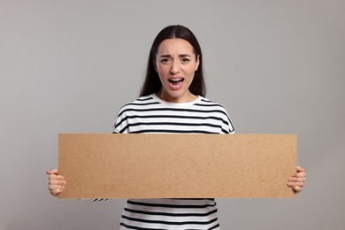 Photo of Angry young woman holding blank cardboard banner on light grey background, space for text