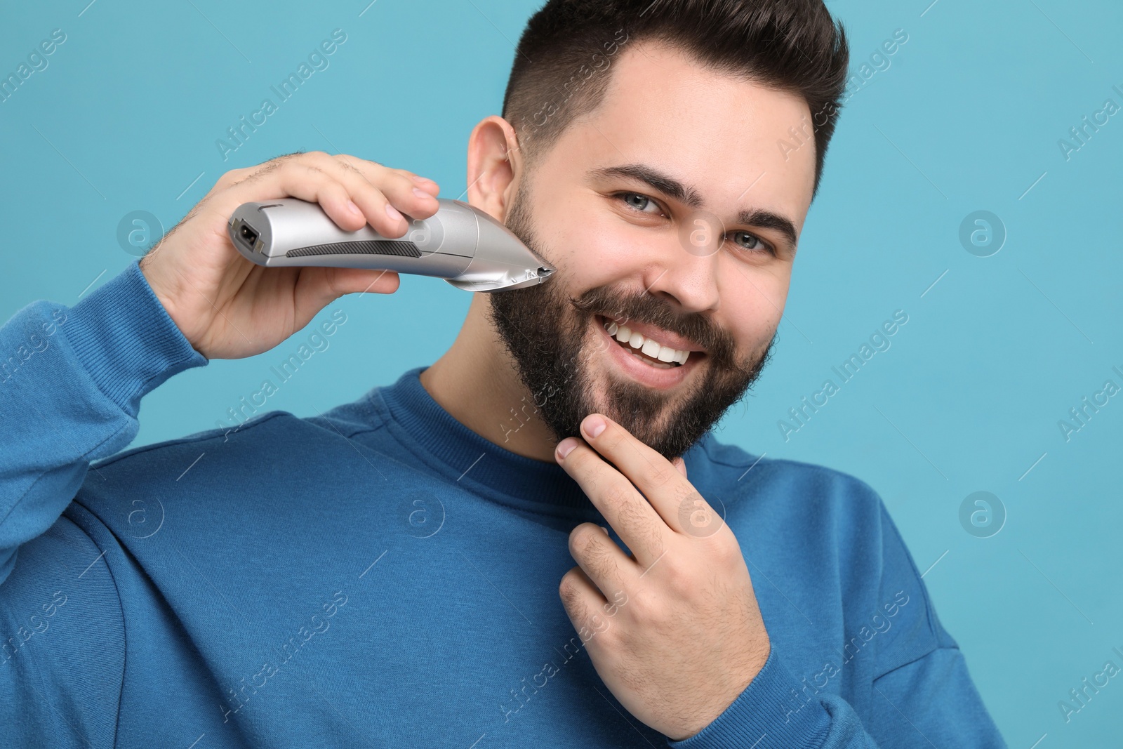 Photo of Handsome young man trimming beard on light blue background