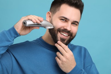 Handsome young man trimming beard on light blue background