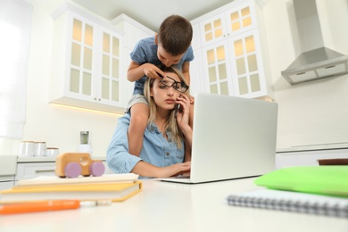 Photo of Little boy bothering mother at work in kitchen. Home office concept