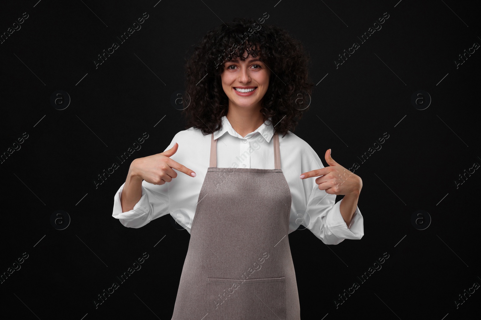 Photo of Happy woman pointing at kitchen apron on black background. Mockup for design