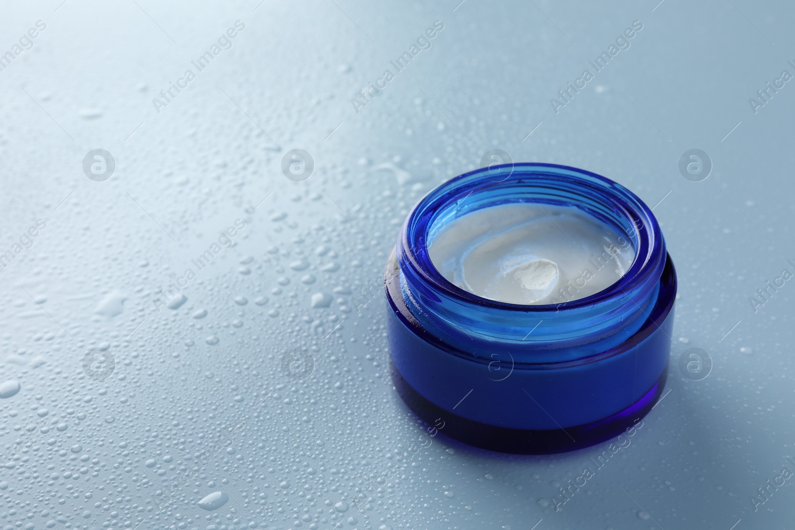 Photo of Moisturizing cream in open jar on light blue background with water drops, closeup. Space for text