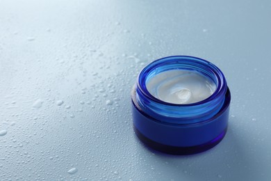 Moisturizing cream in open jar on light blue background with water drops, closeup. Space for text